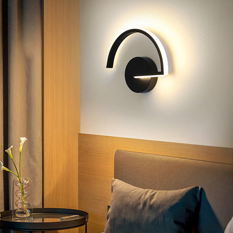 Black LED wall sconce with curved light bar and round backplate, warm glow on wall.