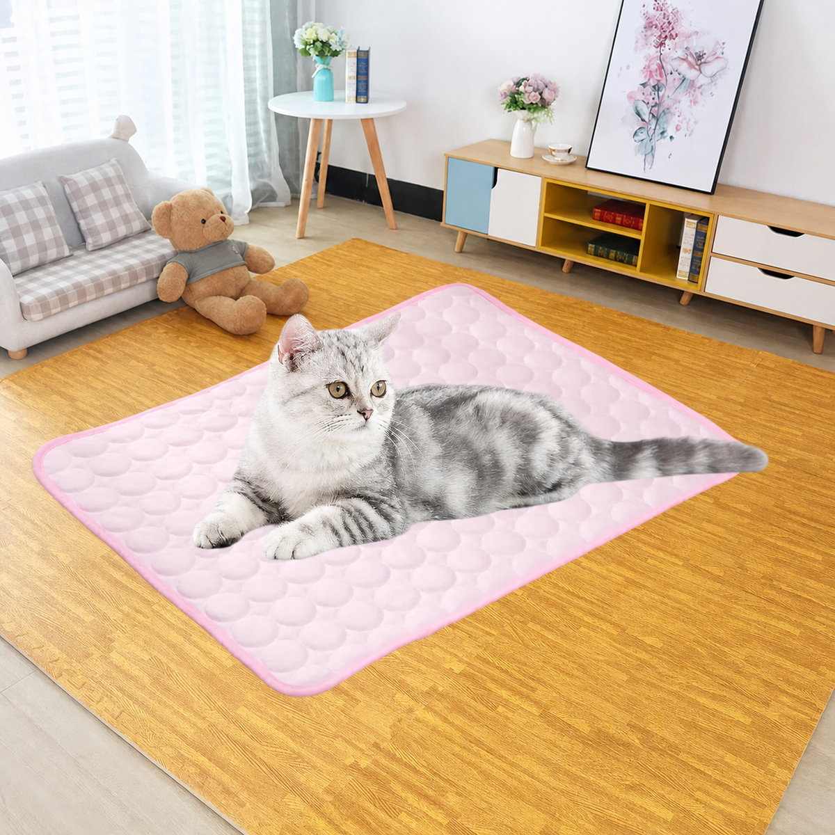 A gray and white cat lies on a pink quilted pet mat placed on a wood-look floor in a bright living room with a sofa, side table, flowers, and storage cabinet.