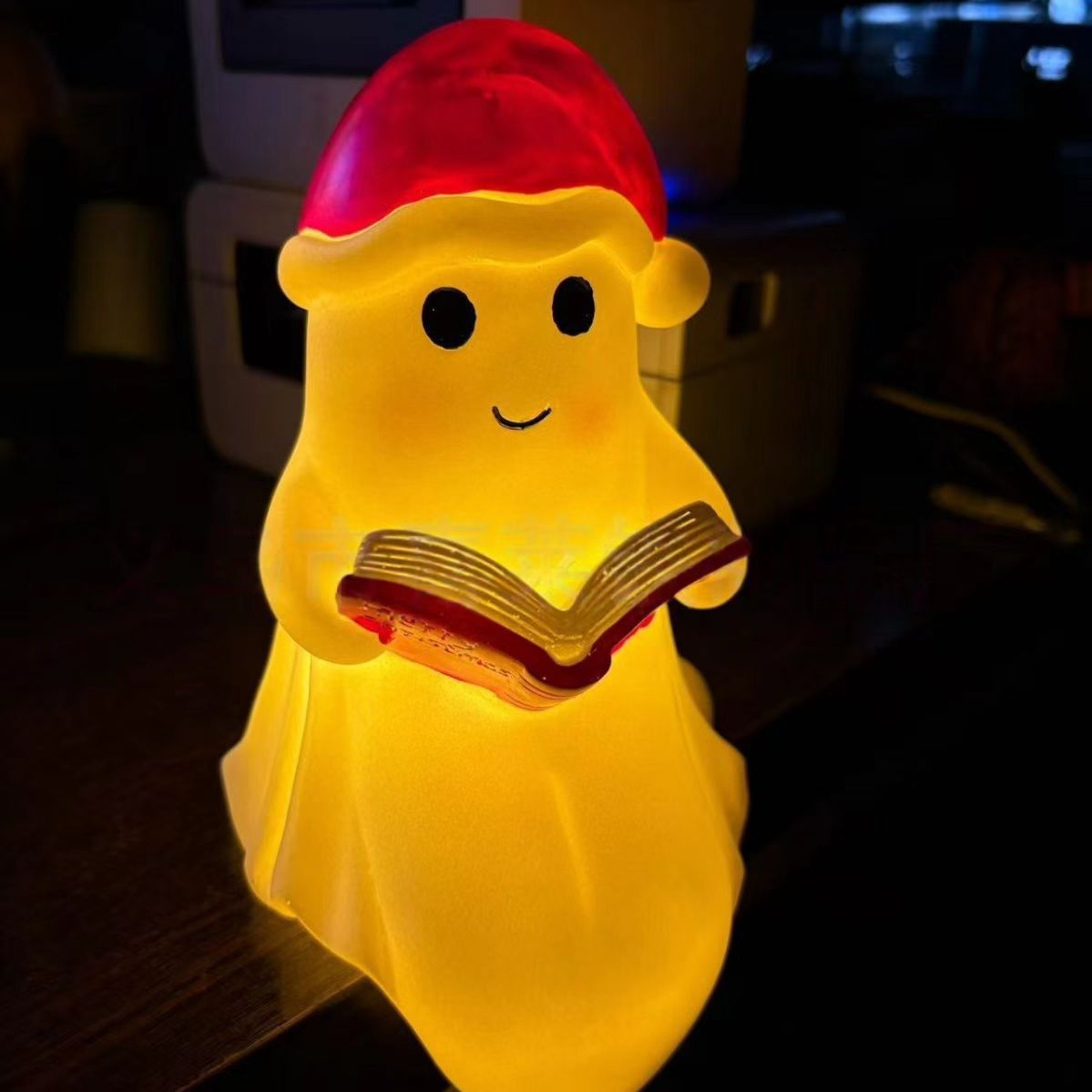 Yellow ghost night light holding an open book with red cap, soft warm glow