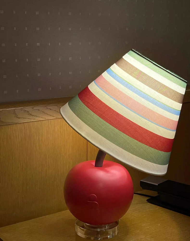 Apple-shaped bedside lamp with striped fabric shade and red base on a wooden surface.