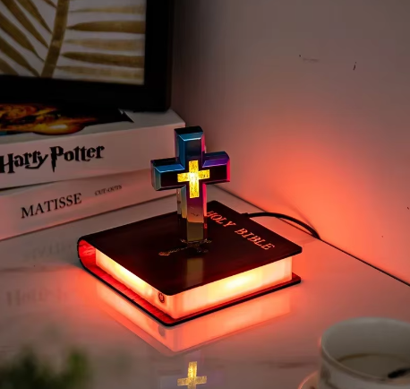 Decorative cross light on a book with a warm glow, placed on a surface with books and a cup in the background.