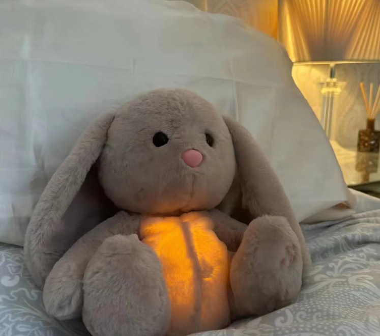 Grey bunny plush music toy on bed with glowing belly