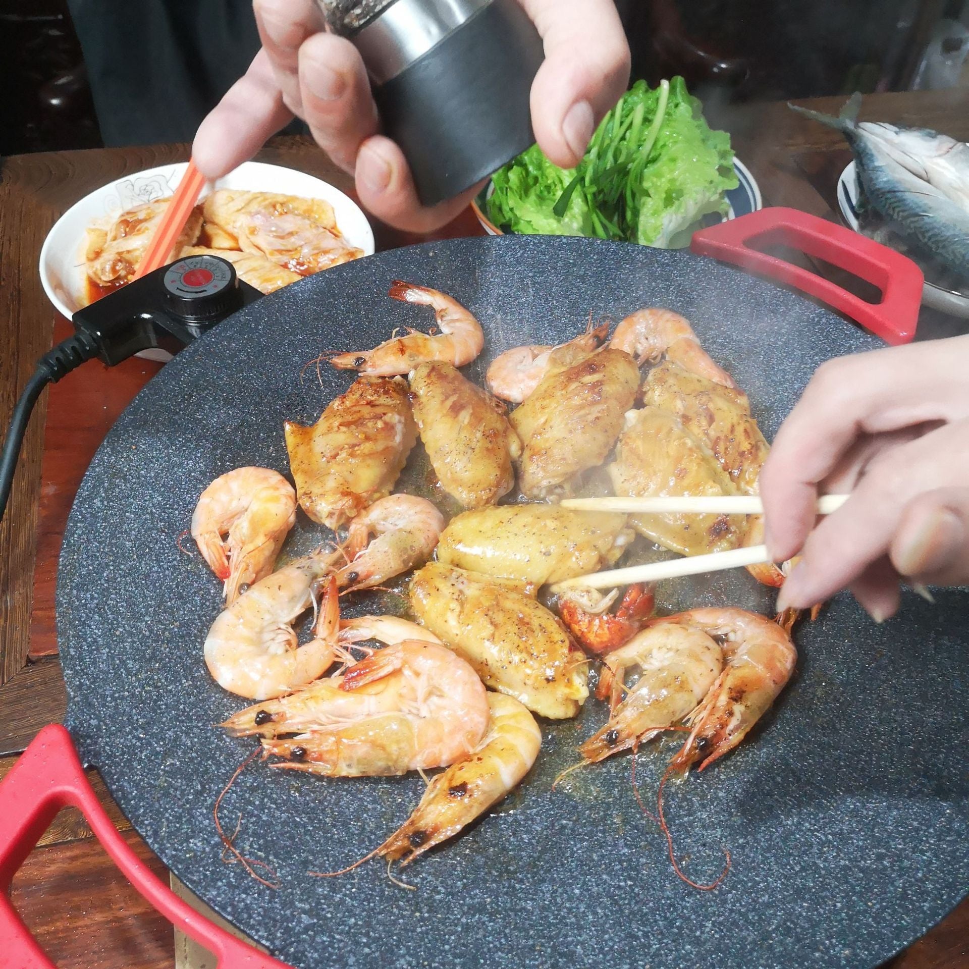 Round electric non-stick grill tray with red handles, cooking surface with assorted meats